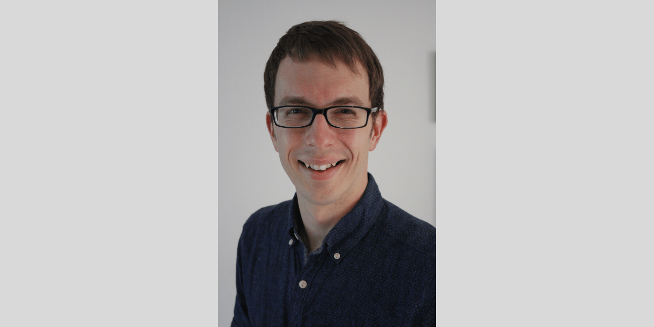A portrait of Tim Hudson, a white man with short dark hair and glasses, smiling at the camera. He wears a navy shirt. The background is grey